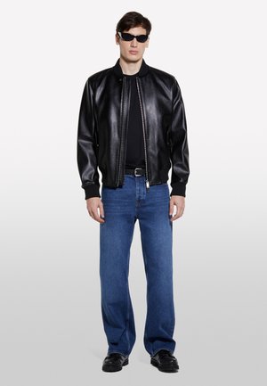 Veste bomber en cuir noir avec des poignets côtelés, t-shirt noir, jeans droit bleu clair, et chaussures noires, portés avec des lunettes de soleil noires.