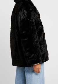 Chaqueta de pelaje sintético negra con un corte holgado, cuello alto y superficie texturizada, llevada sobre unos pantalones vaqueros de denim azul claro.