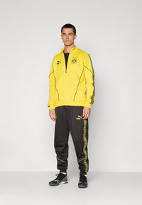 Gul zip-up jakke med sorte accenter og logo, parret med sorte sweatpants med gule sidestripes og logo. Letvægtsstof.