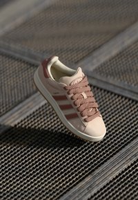 Světlou růžovou tenisku s béžovými detaily, texturovaným povrchem, zavazováním na tkaničky, třemi hnědými proužky na straně a gumovou podrážkou.