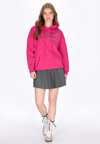 Sudadera con capucha rosa con bolsillo delantero y texto brillante, combinada con una falda plisada gris y zapatillas plateadas con cordones negros y calcetines de malla.
