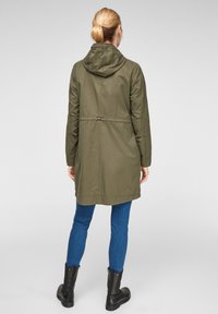 Una donna con capelli biondi raccolti in un bun indossa un cappotto con cappuccio verde oliva, jeans blu e stivali di pelle nera, in piedi su uno sfondo chiaro.