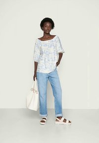 Top claro de ombro à mostra com efeito tie-dye e mangas bufantes, combinado com jeans azuis de perna reta e sandálias brancas de tiras. Grande mala de mão branca.