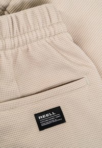 Pantalones cortos texturizados beige con cintura elástica y un bolsillo trasero, que presentan una etiqueta de la marca REELL en negro.