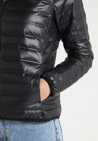 Sort quiltet pufferjakke med en skinnende, glat tekstur, lynlås foran og elastiske manchetter. Har sidelommer og en tætsiddende design.