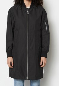 Persona vistiendo una chaqueta bomber negra de longitud a la rodilla con puños acanalados, bolsillos con cremallera en el frente y en las mangas, y un cierre de cremallera plateado, combinada con jeans azules.