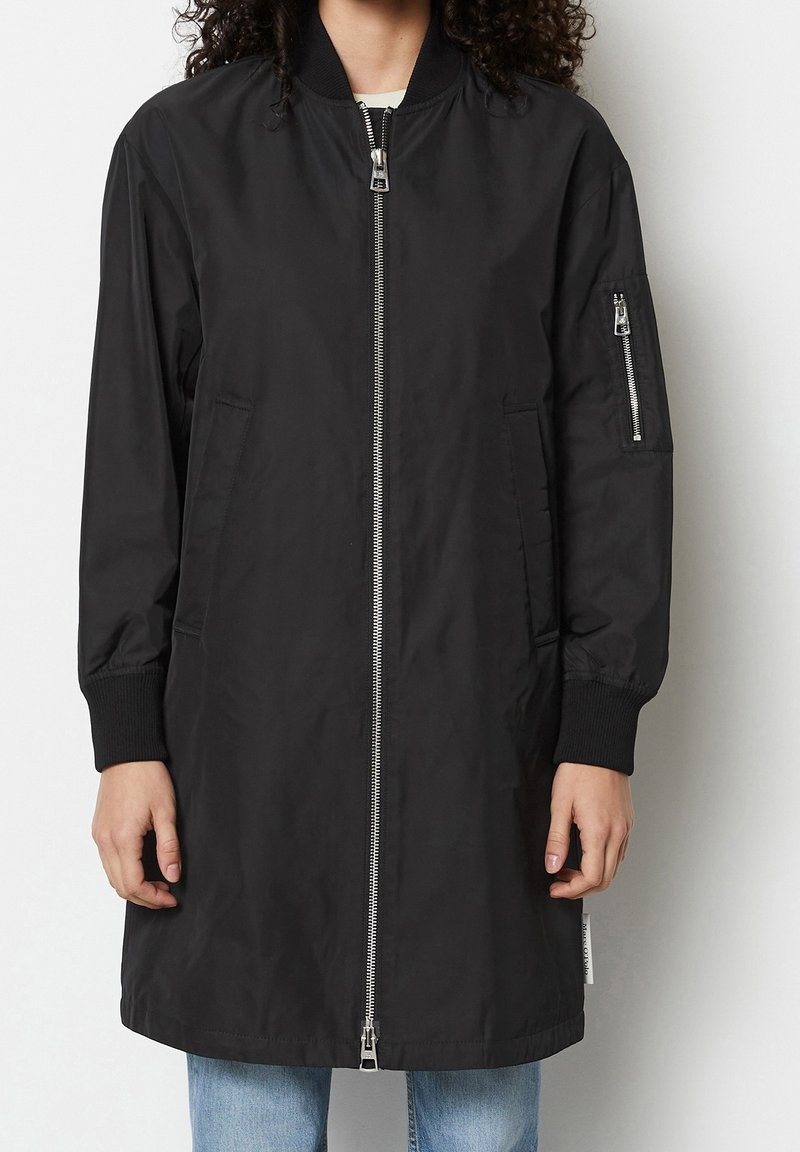 Persona vistiendo una chaqueta bomber negra de longitud a la rodilla con puños acanalados, bolsillos con cremallera en el frente y en las mangas, y un cierre de cremallera plateado, combinada con jeans azules.