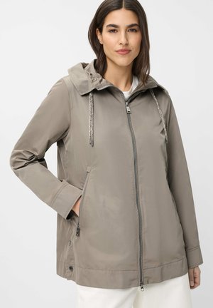 Woman wearing a taupe zip-up hooded jacket with drawstrings, long sleeves, and side zippered pockets, paired with white pants.