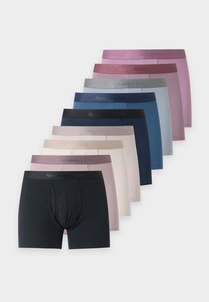 Homme athletic debout pieds nus en boxer noir, regardant sur le côté sur un fond blanc uni.
