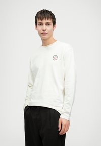 Junger Mann, der ein weißes Langarmhemd mit einem kleinen Globus-Logo und Text trägt, kombiniert mit schwarzen Hosen, steht vor einem einfarbigen Hintergrund.