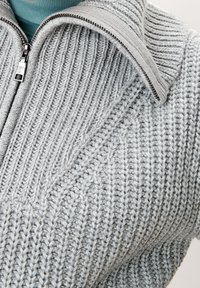 Grå randig stickad tröja med stående krage, featuring en silverfärgad dragkedja och strukturmönster, som framhäver en mysig design.