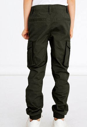 Enfant portant un pantalon cargo vert foncé avec des poignets élastiques et des poches à rabats, baskets blanches, debout sur un fond blanc.
