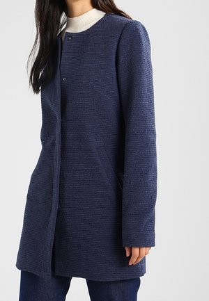 Femme portant un manteau texturé bleu marine à manches longues par-dessus un pull à col roulé blanc, debout devant un fond clair uni.