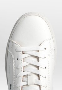 Baskets blanches avec des lacets blancs noués en motif croisé, montrant des coutures détaillées et une texture de cuir lisse sur le dessus.