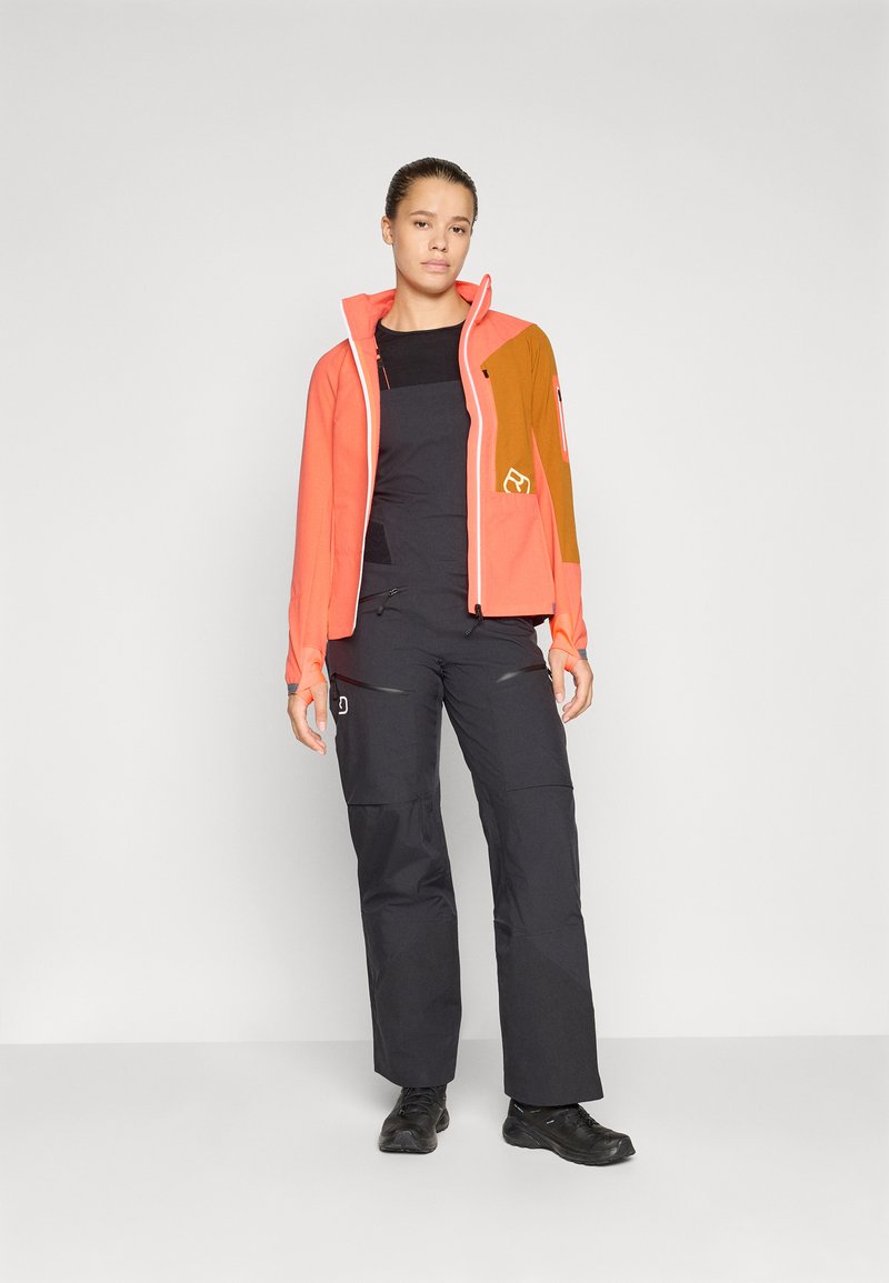 Orange- und braune Zip-Jacke mit kontrastierenden Einsätzen, kombiniert mit schwarzen Latzhosen. Dazu werden schwarze Schuhe getragen. Grauer Hintergrund.