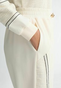 Hoodie texturé crème avec accents noirs, présentant une poche et une taille élastique, mettant en avant un mélange de matériaux et des lignes épurées.