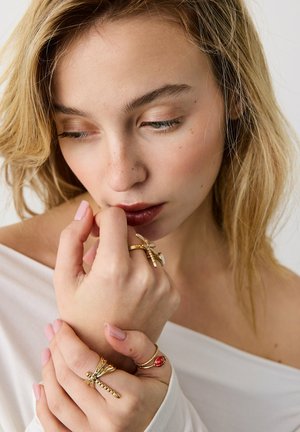 Ung kvinde med lyst hår iført hvidt off-shoulder top, der viser guldregndråberinge og en rød mariehønering på fingrene.