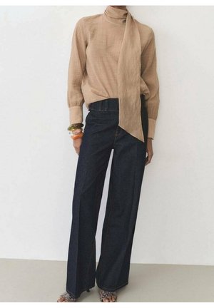 Personne portant une blouse beige à manches longues avec un nœud au cou, un jean large foncé, des talons ouverts à motif serpent et des bracelets colorés au poignet gauche.