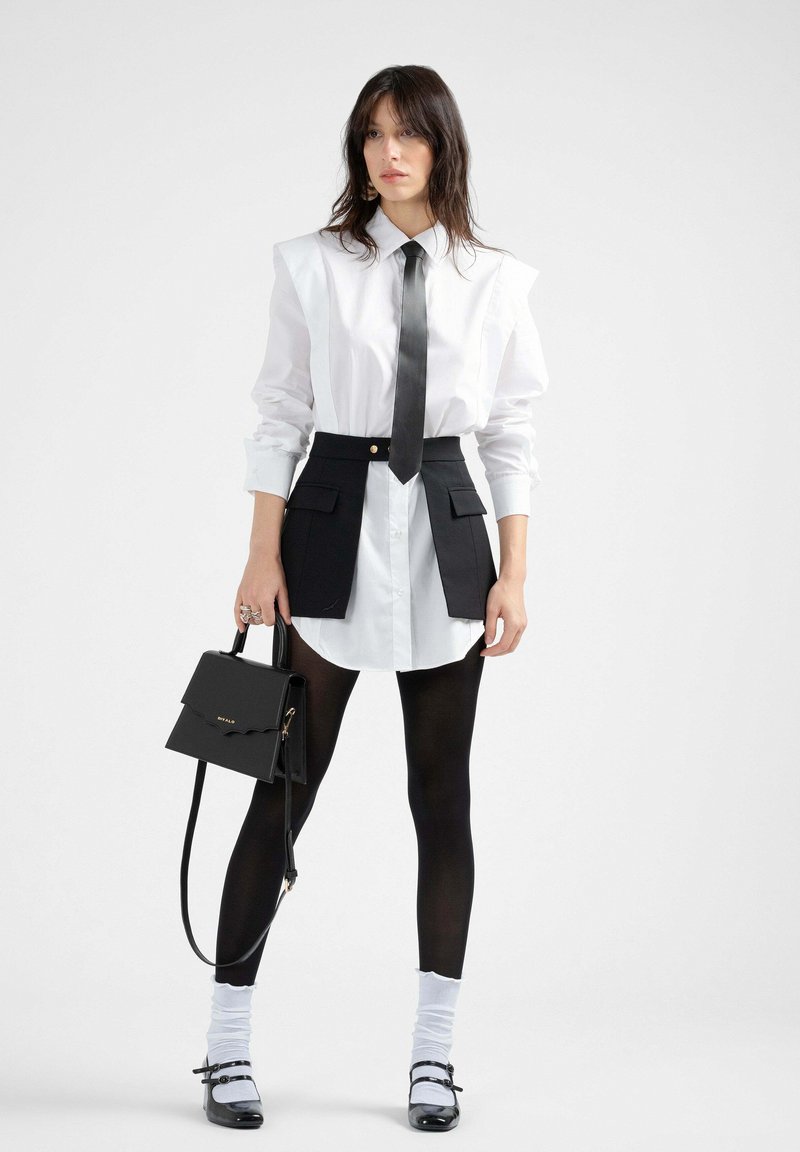 Camicia bianca di grandi dimensioni con design a spalla accentuata, gonna nera in vita con tasche, cravatta nera, borsa nera, calze nere e scarpe in vernice.