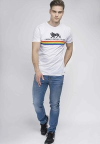 Hvid bomulds T-shirt med et sort løvegrafik og teksten "Lonsdale elsker alle farver," fremhævet med en regnbue-stribe. Båret med blå jeans.