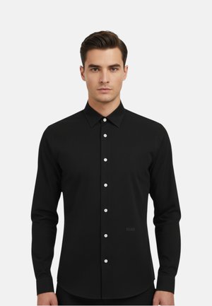 Giovane uomo con capelli corti e scuri che indossa una camicia nera a maniche lunghe con bottoni bianchi su uno sfondo semplice.