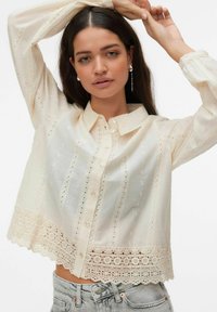 Femme aux longs cheveux bruns portant un chemisier crème orné de dentelle et un jean gris clair, posant avec les bras levés au-dessus de la tête.