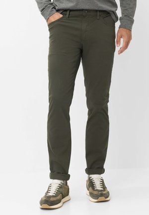 Homme portant un pantalon vert olive roulé aux chevilles avec des chaussettes blanches et des baskets vertes et beige, chemise grise à manches longues partiellement visible.