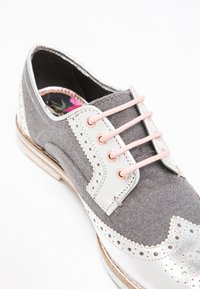 Chaussure en feutre gris à lacets avec des accents en cuir argenté, des lacets roses, des détails brogue et une semelle intérieure à motifs. Semelle plate en bois avec un léger talon.