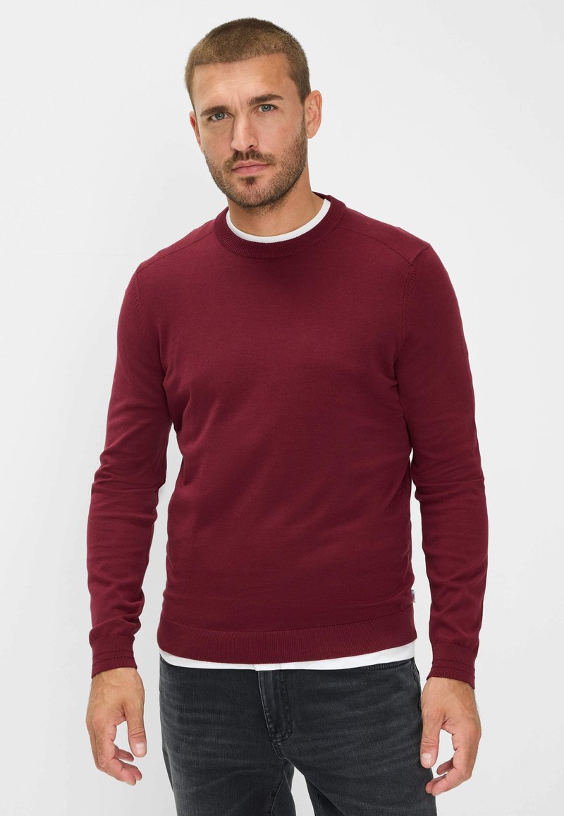 Burgunderfarbener Strickpullover mit langen Ärmeln, rundem Halsausschnitt und gerippten Bündchen. Getragen über einem weißen Hemd mit dunklen Jeans.