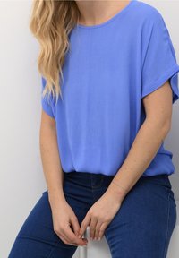 Blå top med korte ærmer i blødt stof, med afslappet pasform og rund halsudskæring, kombineret med mørke denimjeans. Glat tekstur med diskret glans.