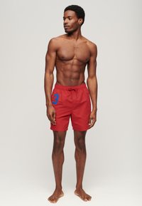 Athletischer Mann steht barfuß in roten Badeshorts mit blauer Nummer 3 und blickt nach links vor einem schlichten hellen Hintergrund.