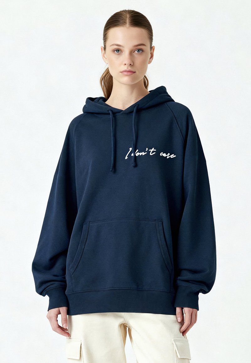 Sweat à capuche bleu marine avec une poche avant, coupe ample, et texte blanc "Je m'en fiche" sur la poitrine. Tissu doux et poignets côtelés.