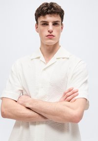 Νέος άνδρας με κοντά, σγουρά μαλλιά, που φοράει λευκό, υφασμάτινο πουκάμισο με κοντά μανίκια, στέκεται με σταυρωμένα τα χέρια μπροστά σε απλό φόντο.