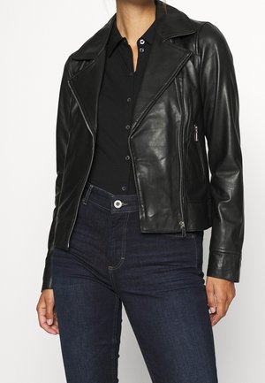 Vrouw die een nauwsluitend zwart leren bikerjack draagt over een zwart overhemd met knopen en donkerblauwe spijkerbroek.