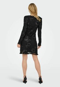 Czarna sukienka z długim rękawem ozdobiona fakturowanym, kwiatowym haftem, sięgająca do połowy uda, z karbowanym dołem i przezroczystą nakładką. Noszona z czarnymi butami na wysokim obcasie.