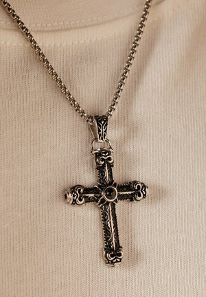 Pendentif en croix argenté avec des motifs complexes, accompagné d'une chaîne texturée. Le pendentif présente une pierre noire centrale.