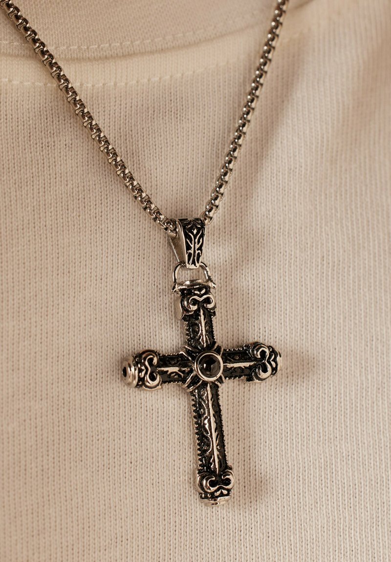 Zilverkleurige kruis hanger met complexe ontwerpen, vergezeld van een gestructureerde ketting. De hanger heeft een centrale zwarte steen.