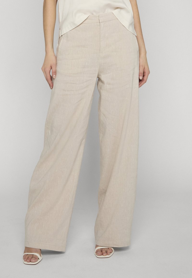 Pantalon beige à jambes larges avec poches avant, porté avec des sandales blanches à lanières et un haut blanc rentré.