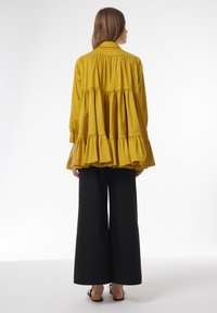 Blusa amarilla mostaza de capas con cuello y dobladillo con volantes, combinada con pantalones negros de pierna ancha y sandalias planas negras.