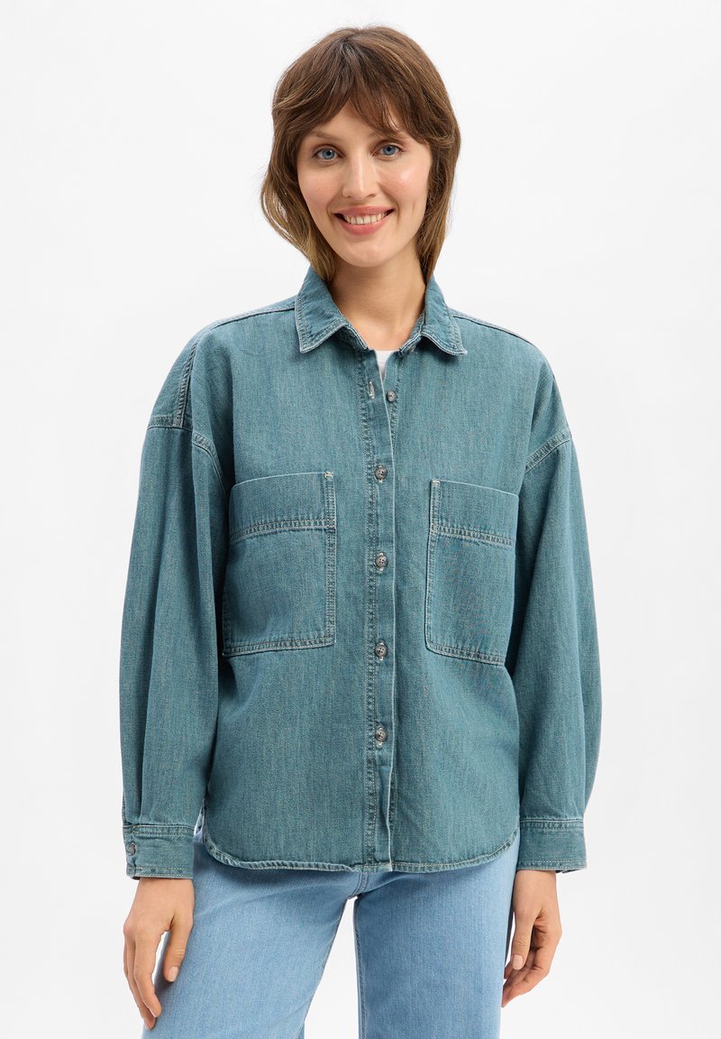 Chemise en denim turquoise avec un devant à boutons, deux grandes poches sur la poitrine et des manches longues. Coupe décontractée avec une silhouette légèrement oversize.