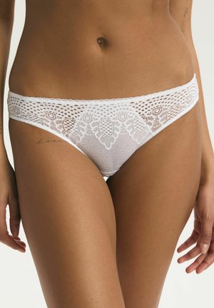 Žena oblečená v bielom čipkovanom bikinovom spodnom prádle s kvetinovými a sieťovanými vzormi, ukazujúca strednú časť tela a hornú časť stehien.