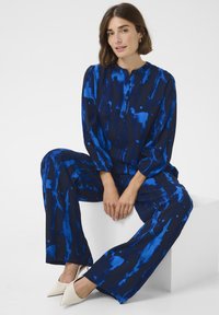 Sininen kahden osan asu, jossa on abstrakteja sinisiä kuvioita. Ominaisuuksiin kuuluu napitettu kaulus ja rento malli, joka on yhdistetty teräväkärkisiin valkoisiin korkokenkiin.