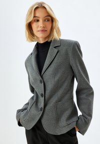 Blazer grigio in misto lana con design sartoriale, rever a bottoni, due tasche frontali e bottoni neri. Texture liscia, silhouette aderente.