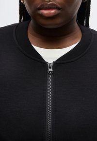 Κοντινό πλάνο ενός ατόμου που φοράει μαύρο μπουφάν με φερμουάρ πάνω από λευκό πουκάμισο, εμφανίζοντας το φερμουάρ και το κάτω μέρος του προσώπου χωρίς μάτια.