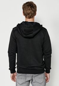 Felpa nera con cappuccio e trama a coste, dotata di tasca a marsupio e polsini elastici. Indossata con jeans grigio chiaro.