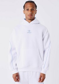 Jonge man met gevlochten haar draagt een witte hoodie en bijpassende sweatpants met lichtblauwe piping, staand tegen een effen witte achtergrond.