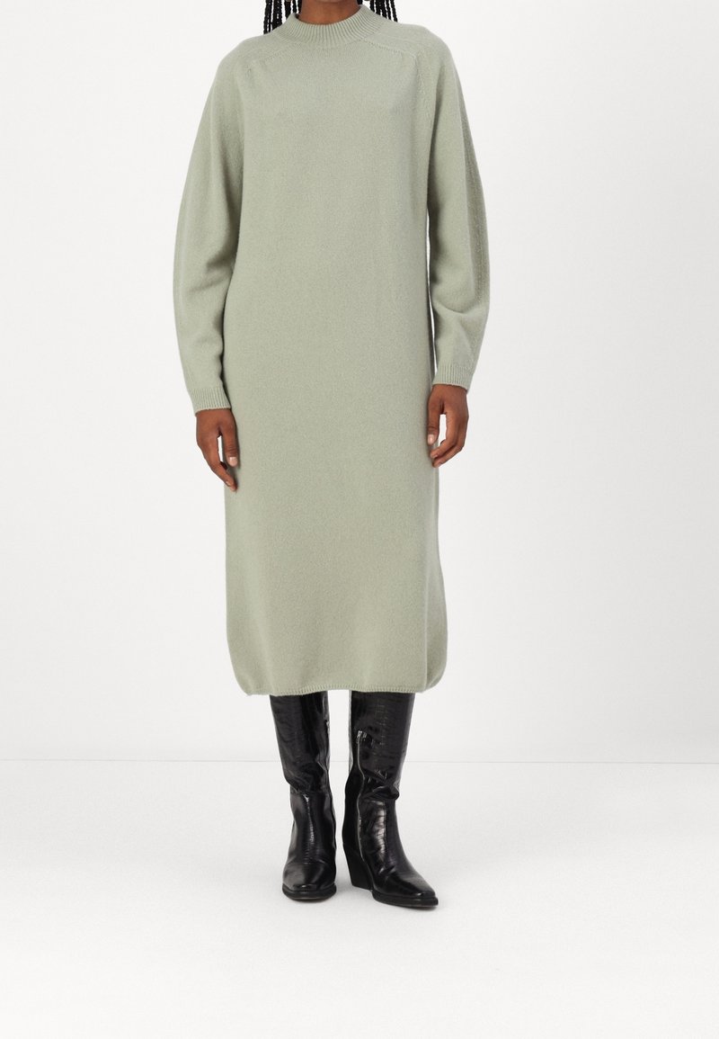 Femme portant une longue robe pull tricotée vert clair associée à des bottes en cuir noires hautes jusqu'au genou, se tenant devant un fond blanc.