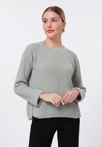 Harmaa ribattu neule, jossa on pitkät hihat, teksturoidut pitsikoristeet olkapäissä, pyöreä pääntie ja etupuolella halkiot.