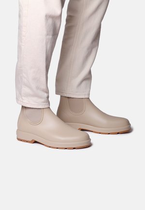 Béžové nepremokavé členkové topánky s elastickými bočnými panelmi, gumovým materiálom, textúrovanou podrážkou a svetlohnedou podrážkou, spojené s nohavicami krémovej farby.