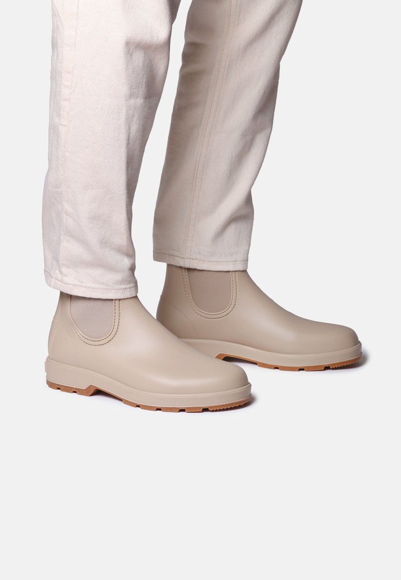 Botas tobilleras impermeables beige con paneles laterales elásticos, material de caucho, suela texturizada y suela exterior de color marrón claro, combinadas con pantalones de color crema.